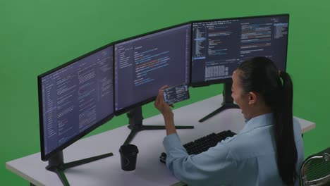 side view of happy asian woman developer looking at code on mobile phone while write code with multiple computer screens in green screen studio