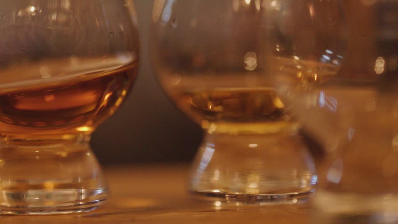 Two whiskey glasses with amber liquid sit on a wooden bar, warm ambient lighting, shallow depth of field, and slow lateral camera movement