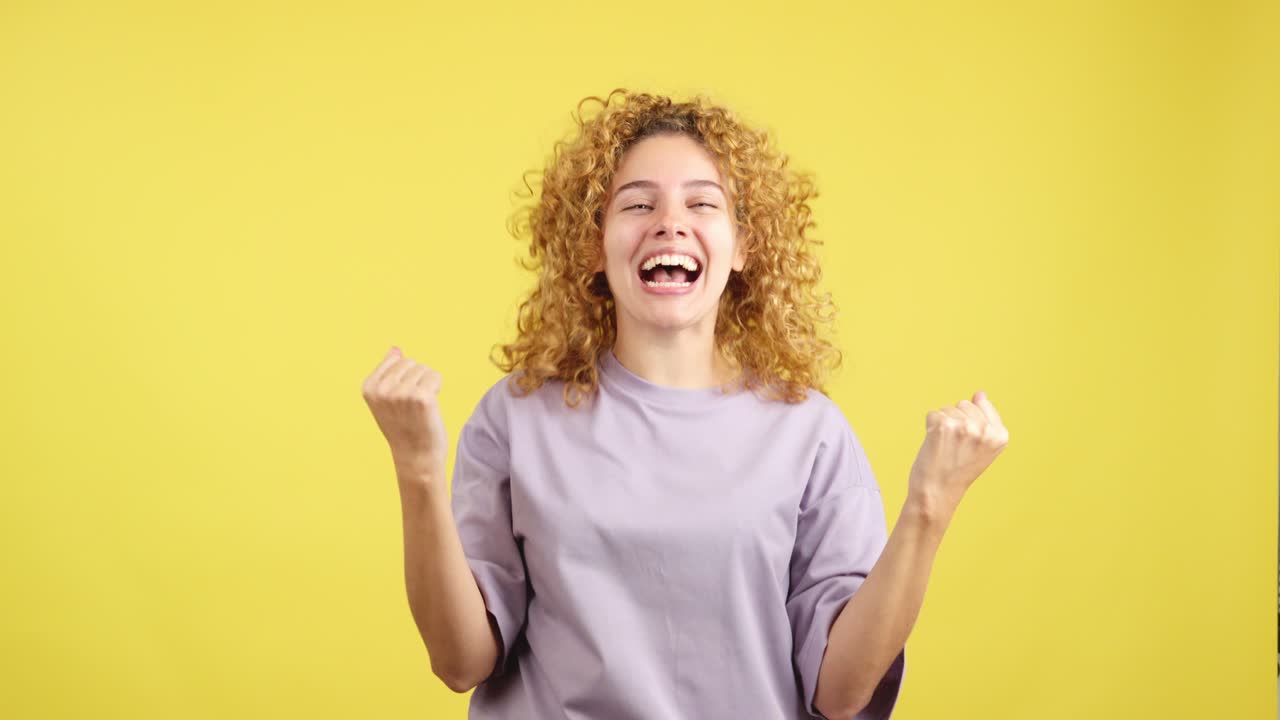 Ecstatic curly-haired woman celebrating success with clenched fists on a vibrant yellow background