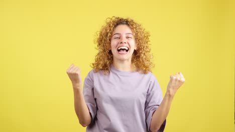 ecstatic curly-haired woman celebrating success with clenched fists on a vibrant yellow background