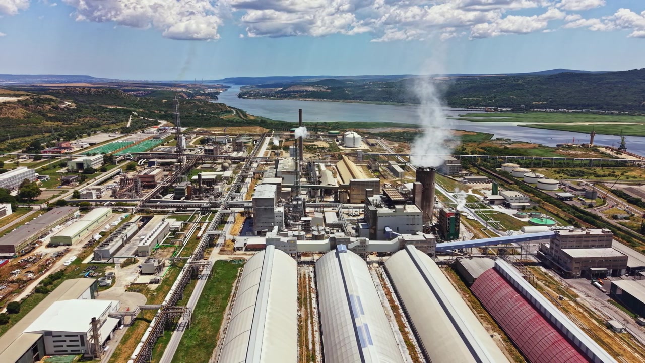 Industrial area near a river with factories, storage buildings, and smoke stacks during daytime with blue sky and clouds in background