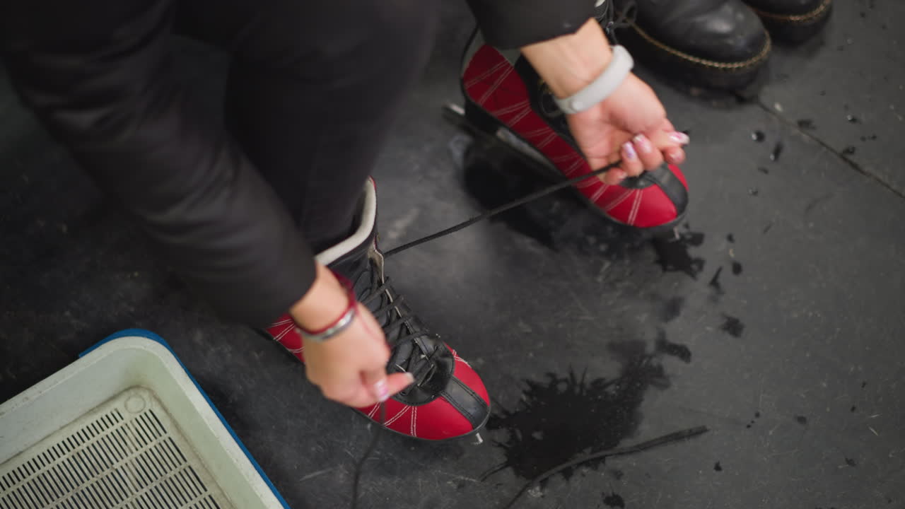Lady putting on red black ice skates while tightening laces on wet floor near bench, preparing for skating session with focus on footwear, blades, water drops, and training atmosphere inside arena