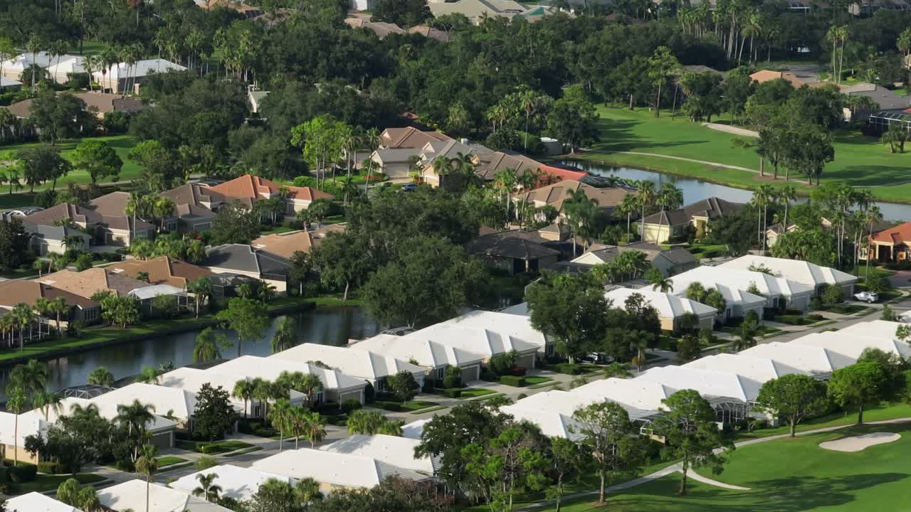 Aerial view of Florida golf community with white-roof homes, palm trees, waterways and green fairways under bright sunshine. Tropical suburban living in USA