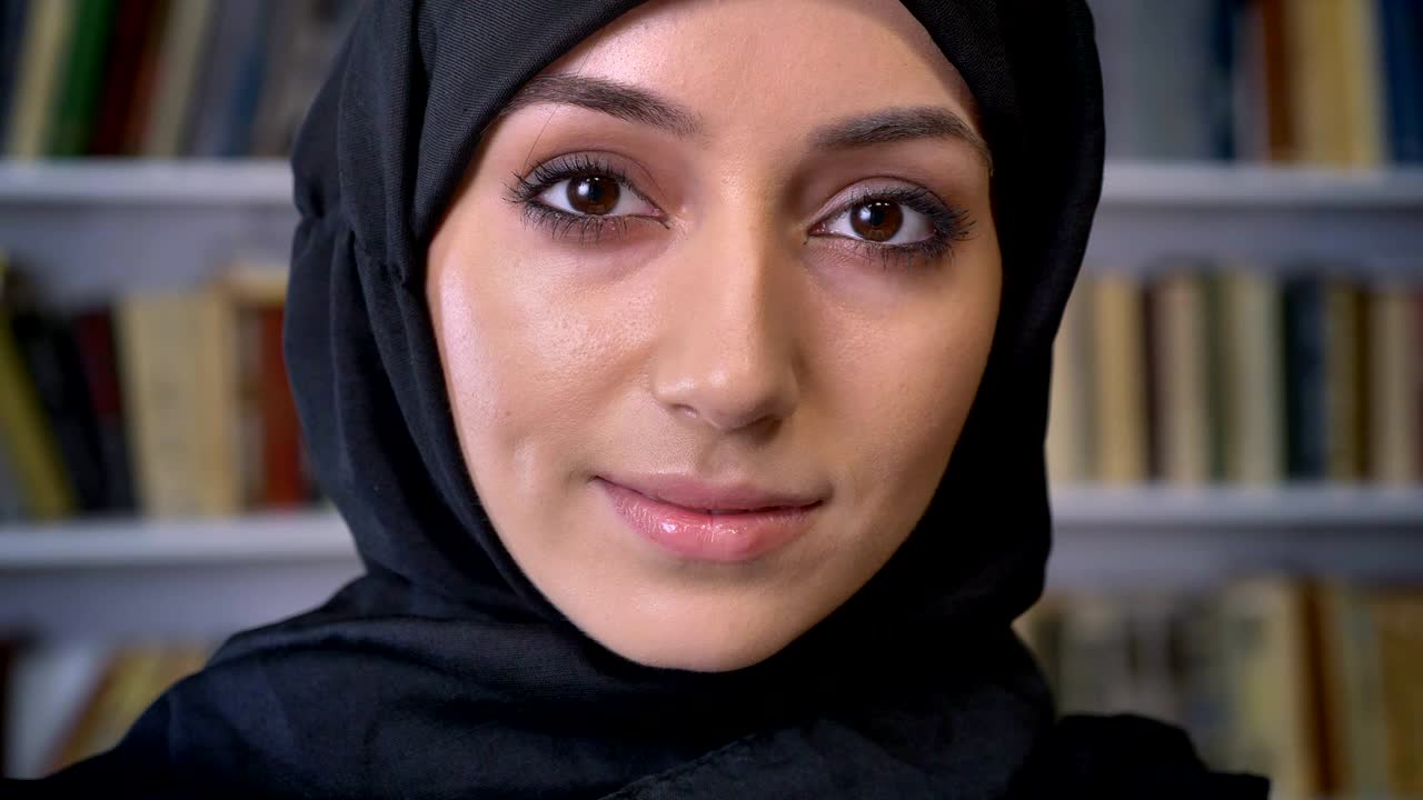 Young beautiful muslim girl in hijab is watching down in book, watching at camera, religious concept, bookshelf on background, blurred background