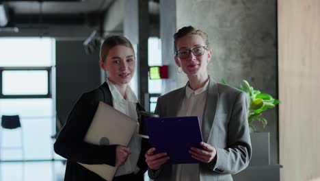 Portrait-of-two-blonde-businesswoman-in-a-business-suit-who-holds-a-laptop-and-tablet-in-her-hands-while-working-in-a-modern-office.-A-blonde-girl-and-her-mentor-a-middle-aged-girl-pose-in-a-modern-office