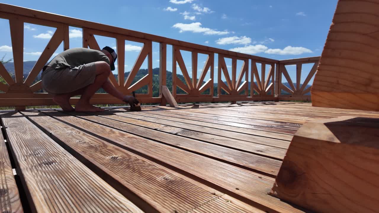 Man oils deck boards in summer sun. Decorative railing, gloves, brush, and mountain horizon highlight home renovation and outdoor wood preservation