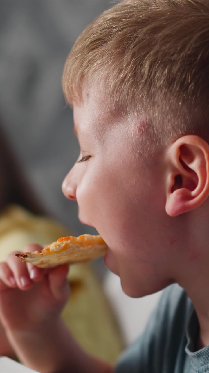 Close up of young boy eating slice of pizza. Boy holds and takes bites from food. Soft lighting highlights his profile as boy enjoys each bite with background remaining out of focus