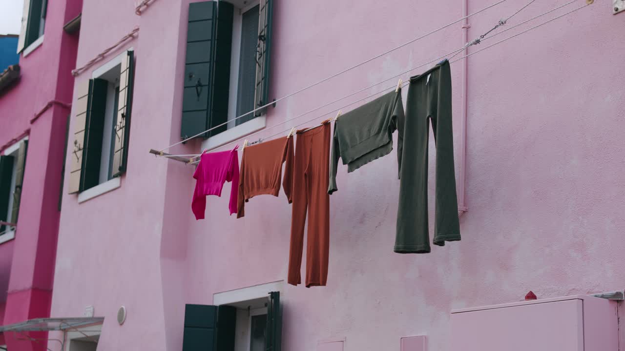 Scena pittoresca dell'Isola di Burano, Venezia, che mostra un edificio rosa pastello con vestiti stesi ad asciugare su uno stendibiancheria, creando un'atmosfera vivace e affascinante.