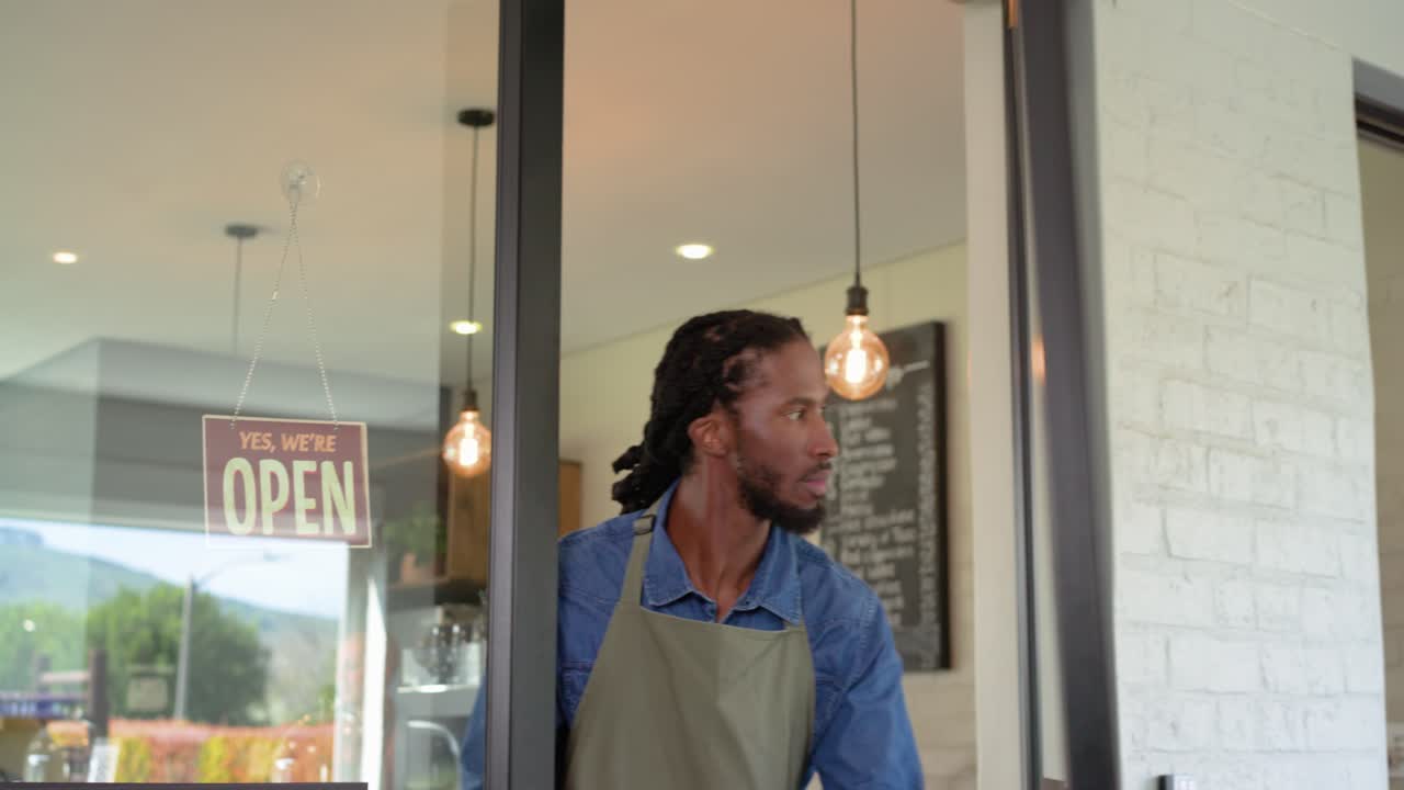 Barista maschio afroamericano che gira il cartello "chiuso", apre la porta ed entra nel caffè per aprire, con spazio per il testo.
