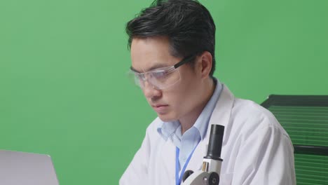 close up of side view of asian man scientist typing on a laptop while working on the table with microscope and test tube in the green screen background laboratory