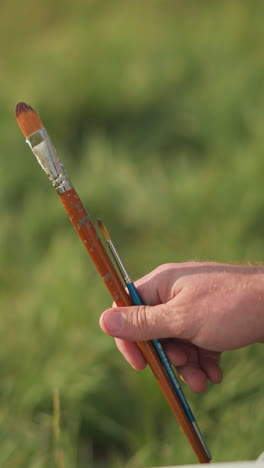a close-up shot of a man's hand holding two paintbrushes one small and one very small against a blurred green background. the scene captures the simplicity and focus on the tools of artistic creation