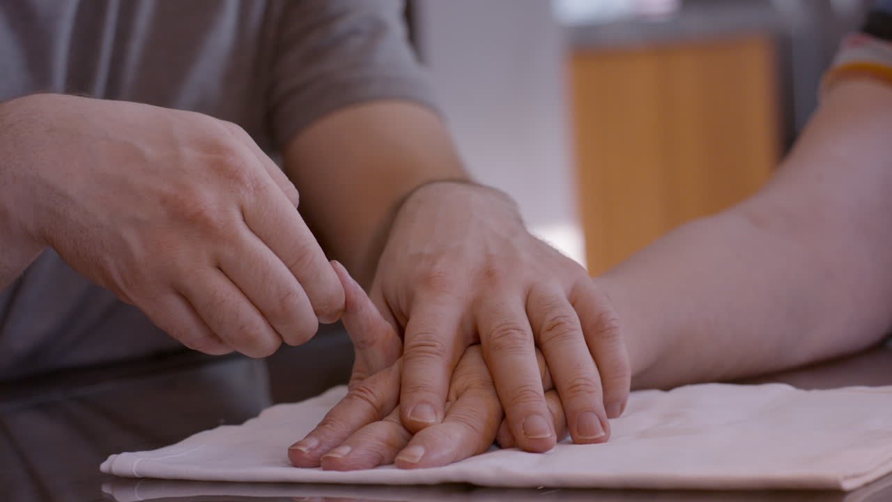 Adult son caregiver assists elderly mother stroke survivor by gently stimulating and mobilizing fingers of her hemiplegic right hand during a therapy session at home kitchen table on cloth pad. 24fps.