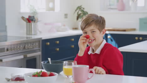 laughing boy wearing school uniform in kitchen putting strawberry on nose as he eats breakfast