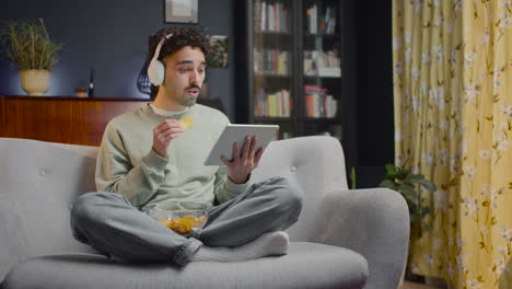 young man watching unexpected movie scene on tablet while eating chips and sitting with crossed legs on couch at home