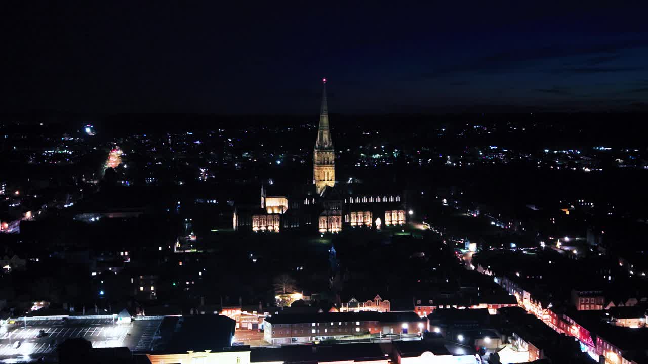 Ripresa aerea con drone della Cattedrale di Salisbury illuminata di notte con le luci della città circostanti nel Wiltshire, Regno Unito, che mostra un chiaro skyline di riferimento, adatto per documentari di viaggio e filmati di apertura.