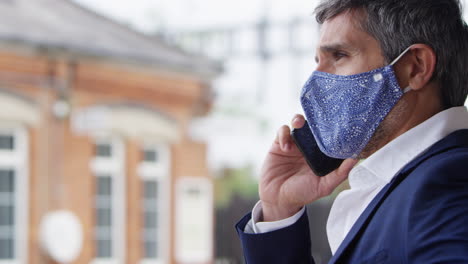 businessman on railway platform talking on mobile phone wearing ppe face mask during health pandemic