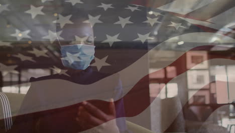 animation of american flag over african american woman in face mask using smartphone