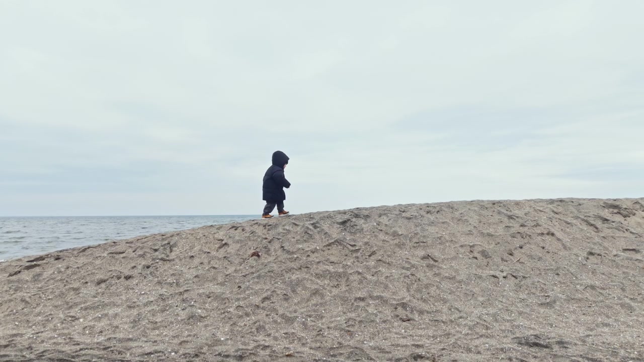 Criança caminha em duna de areia perto da praia sob céu nublado com água ao fundo em clima fresco.