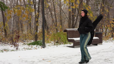 music artist in winter jacket with hands in pockets and guitar on back walking in snowfall, snowy bench and dry trees in background, serene winter landscape