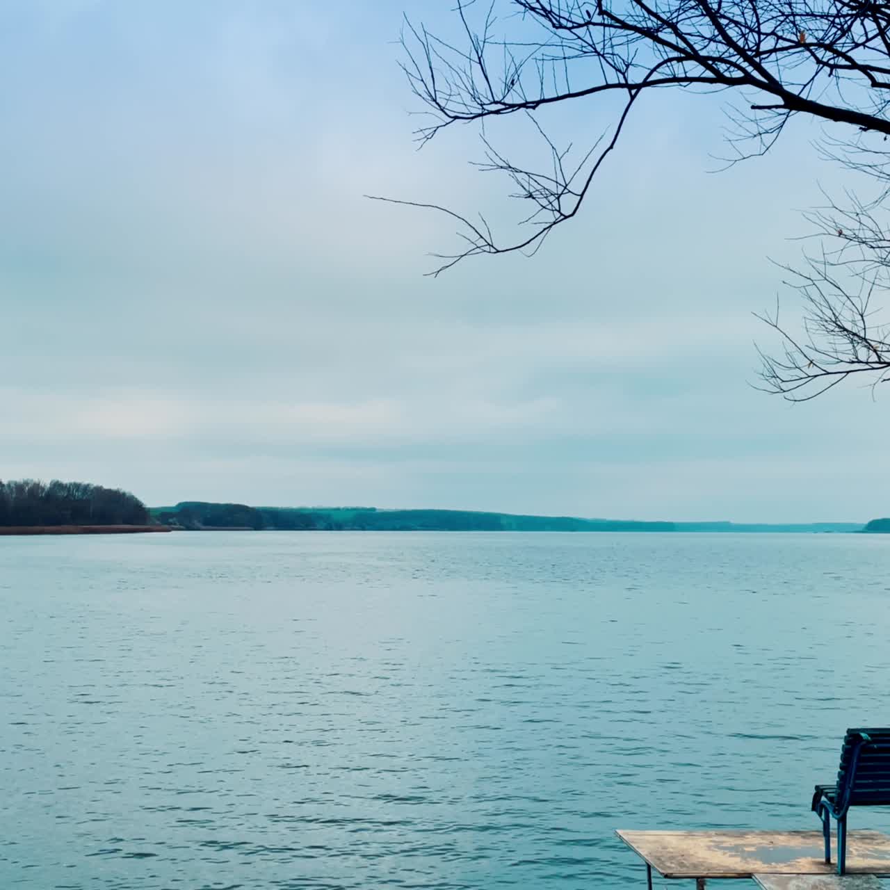 Grey dull day on the river in autumn season. Little footbridge with deckchair on. Cloudy sky and hazy horizon at backdrop