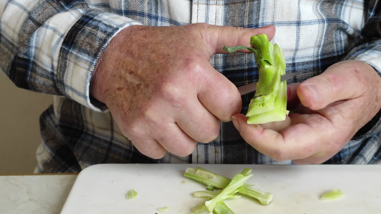 Closeup of male hands peeling broccoli stems over a cutting board while sitting on a table.