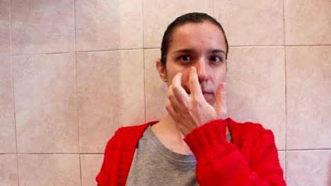 Young-woman-with-acne-touching-her-face-skin-in-bathroom,-medium-close-up