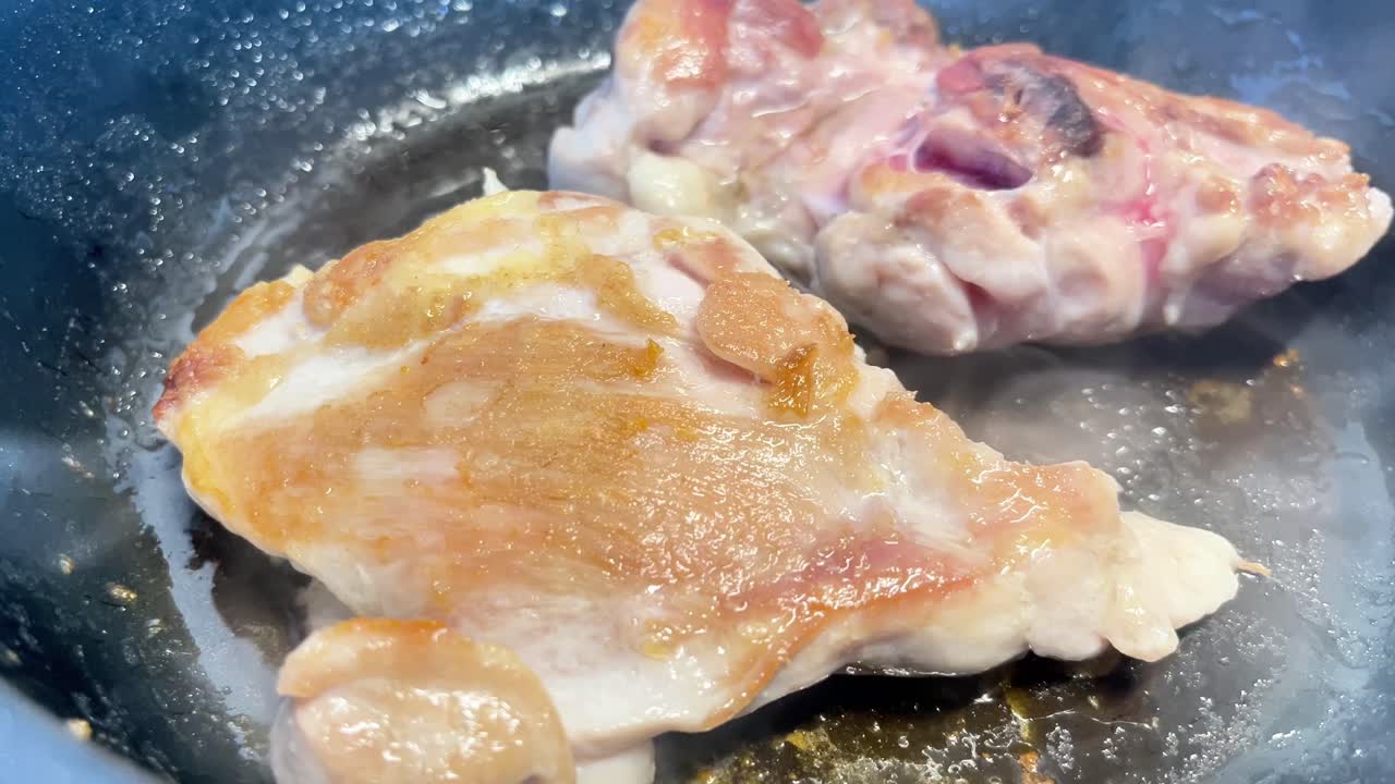 Due pezzi di pollo stanno cuocendo in una padella. Il pollo è dorato e croccante. La padella è nera e il pollo è bianco.