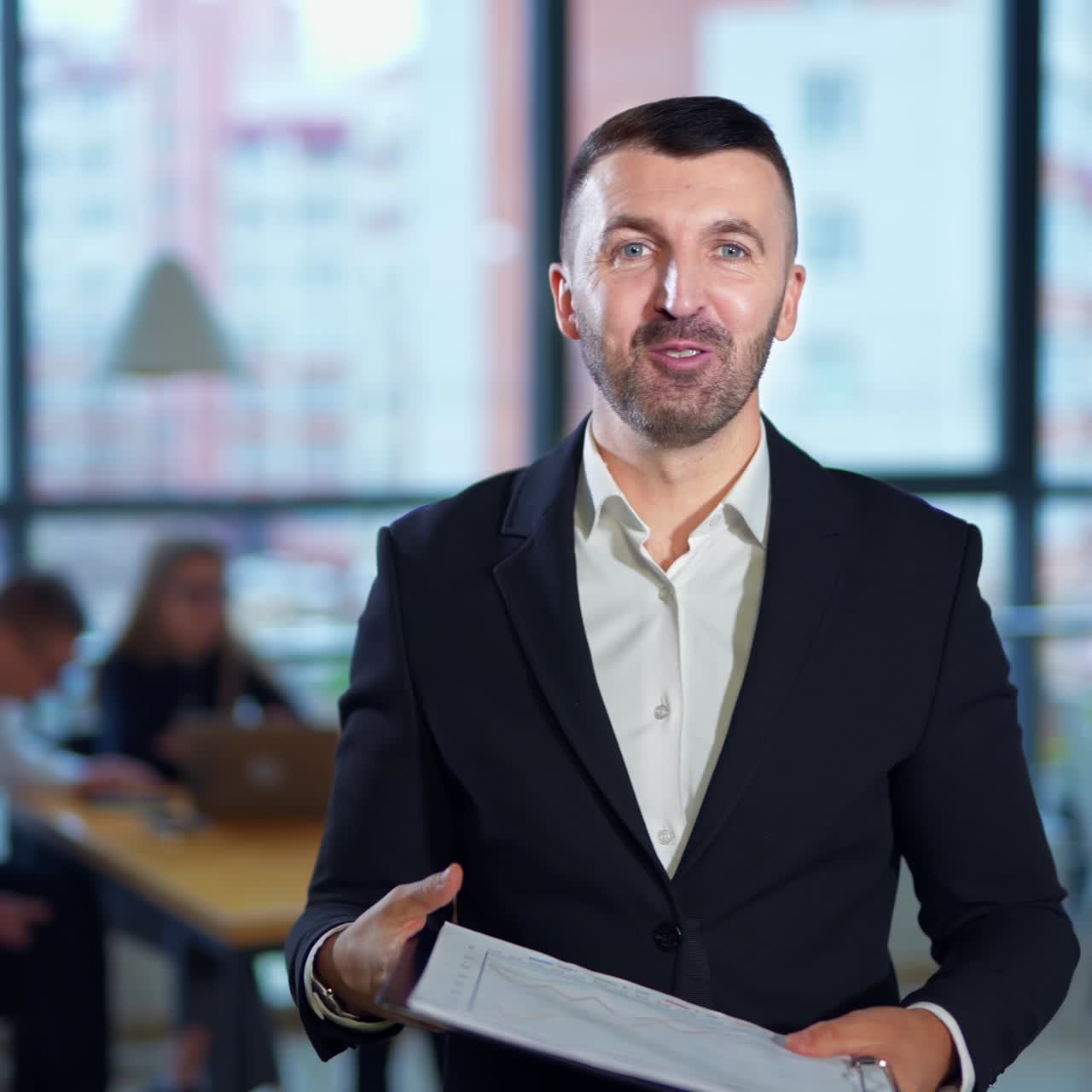 Good-looking energetic bearded man in suit talks to camera holding a folder. Man shows the chart and comments it. Office workers in blur at backdrop
