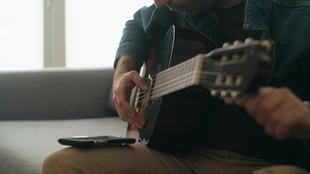 Un uomo con una giacca di jeans seduto su un divano che suona una chitarra acustica classica, usando uno smartphone per gli accordi o per registrare. Scena autentica per contenuti di educazione musicale e benessere.