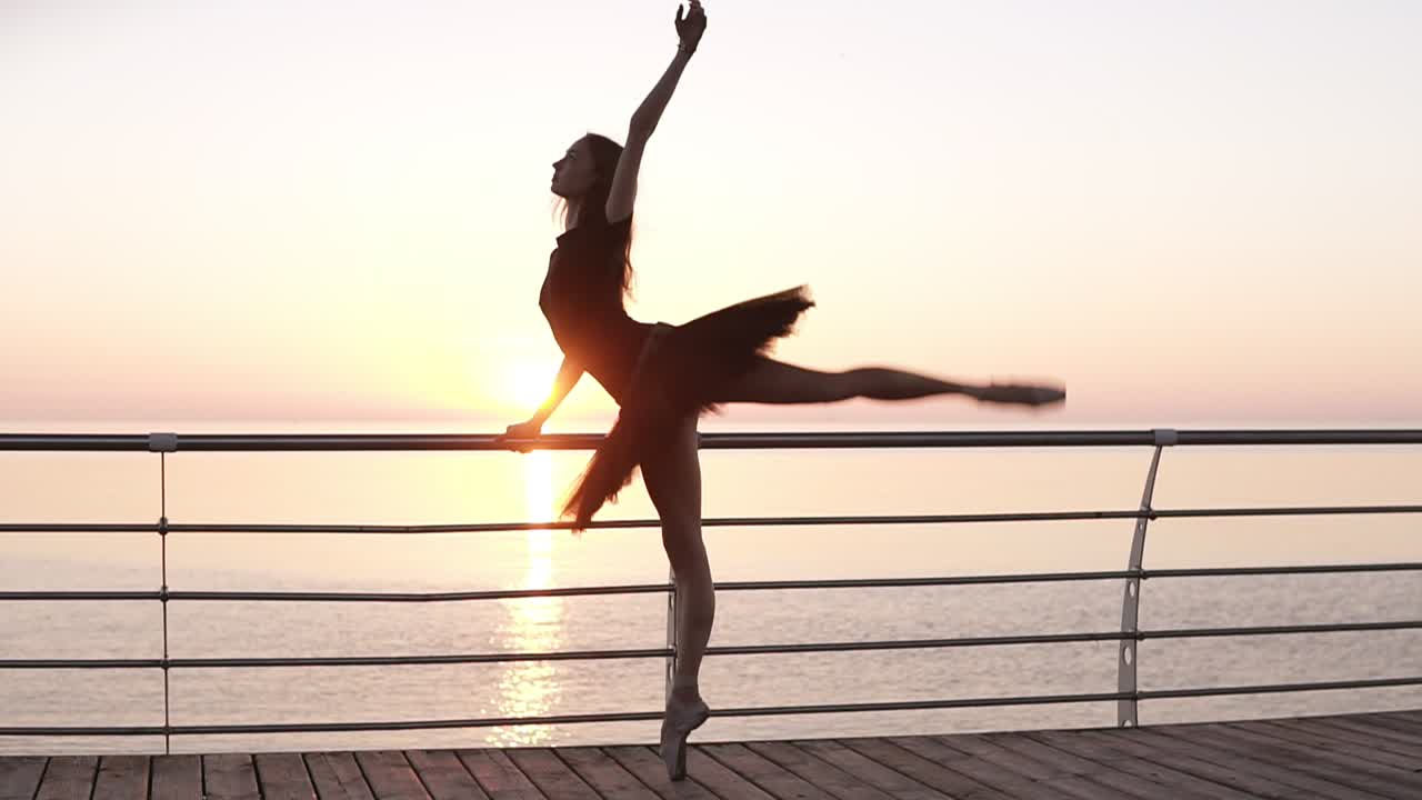 longitud completa. una bailarina de ballet en un tutu negro, haciendo ejercicios, apoyada en una barra cerca del mar. joven bailarina de ballet estira sus piernas en una sentadilla. mañana, amanecer rosado. cámara lenta