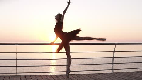 full length. a ballet dancer in a black tutu, doing exercises, leaning on a bar near the sea. young ballet dancer stretches her legs in a squat. morning, pink sunrise. slow motion