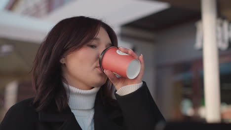 freelancer sips coffee, then gently places cup back on table before turning focus back to laptop, cozy outdoor workspace features wooden chair and flowers in blurred background