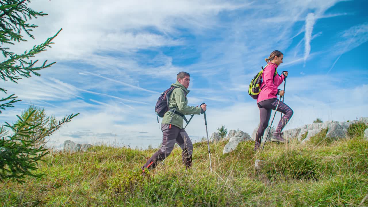 Young Couple Walking Across Rocky Grassland With Trekking Poles. Follow Shot, Slow Motion