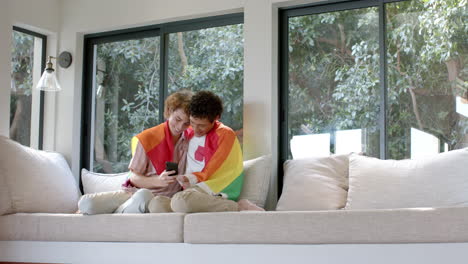 Pareja-Gay-Diversa-Con-Bandera-Lgbt-Arcoiris,-Usando-Un-Teléfono-Inteligente-En-Casa,-Cámara-Lenta,-Espacio-Para-Copiar