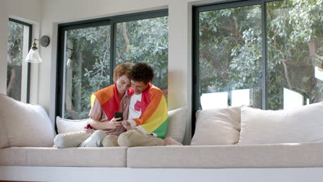 pareja de hombres homosexuales diversos con bandera lgbt arco iris, usando teléfono inteligente en el hogar, cámara lenta, espacio de copia