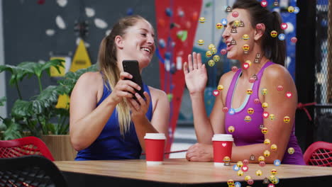Two athletic women sharing smartphone in climbing gym lounge, showing ad icons and graph overlays