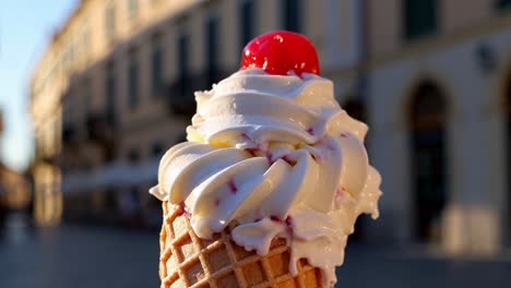 creamy vanilla ice cream with a candied cherry on top melting under the summer sun in a waffle cone, with a blurred city center in the background