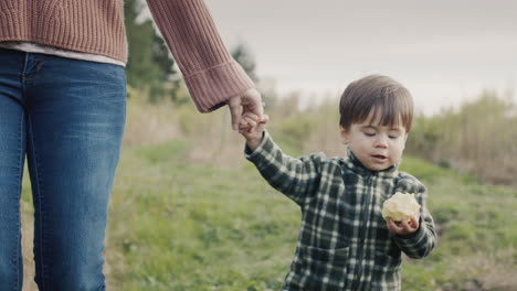 mom leads a little boy by the hand, he eats an apple on the go