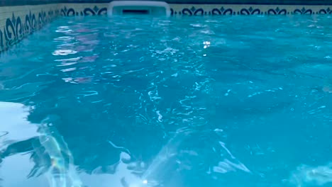 Closeup-of-open-air-clean-swimming-pool-with-rippling-water-surface-and-glimmering-sunlight-rays-and-skimmer-in-the-background-in-slow-motion