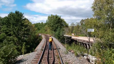 dolly in drone shot of a man with a yellow backpack on walking on a train rail surrounded by pine trees in canada