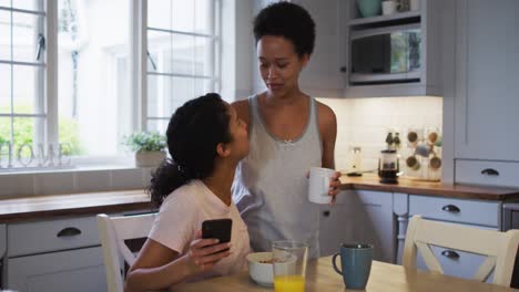 Mixed-race-lesbian-couple-hugging-and-drinking-coffee-in-kitchen