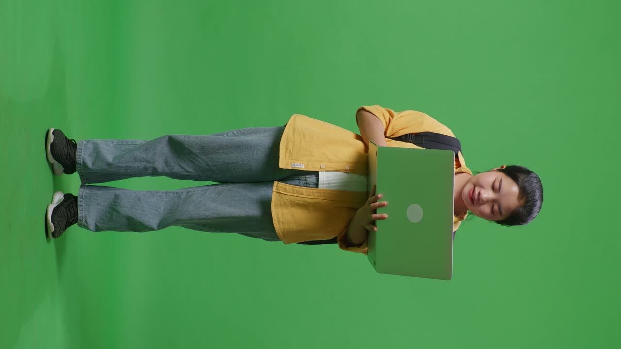 cuerpo lleno de estudiante mujer asiática con una mochila sonriendo y escribiendo en una computadora portátil mientras estaba de pie en el estudio de fondo de la pantalla verde