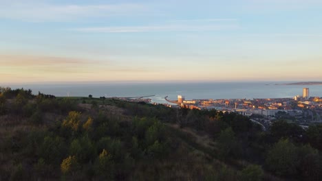 Vista-Aérea-En-Aumento-De-Las-Colinas-Y-La-Ciudad-Al-Amanecer-Con-Edificios-Y-Puerto-Deportivo