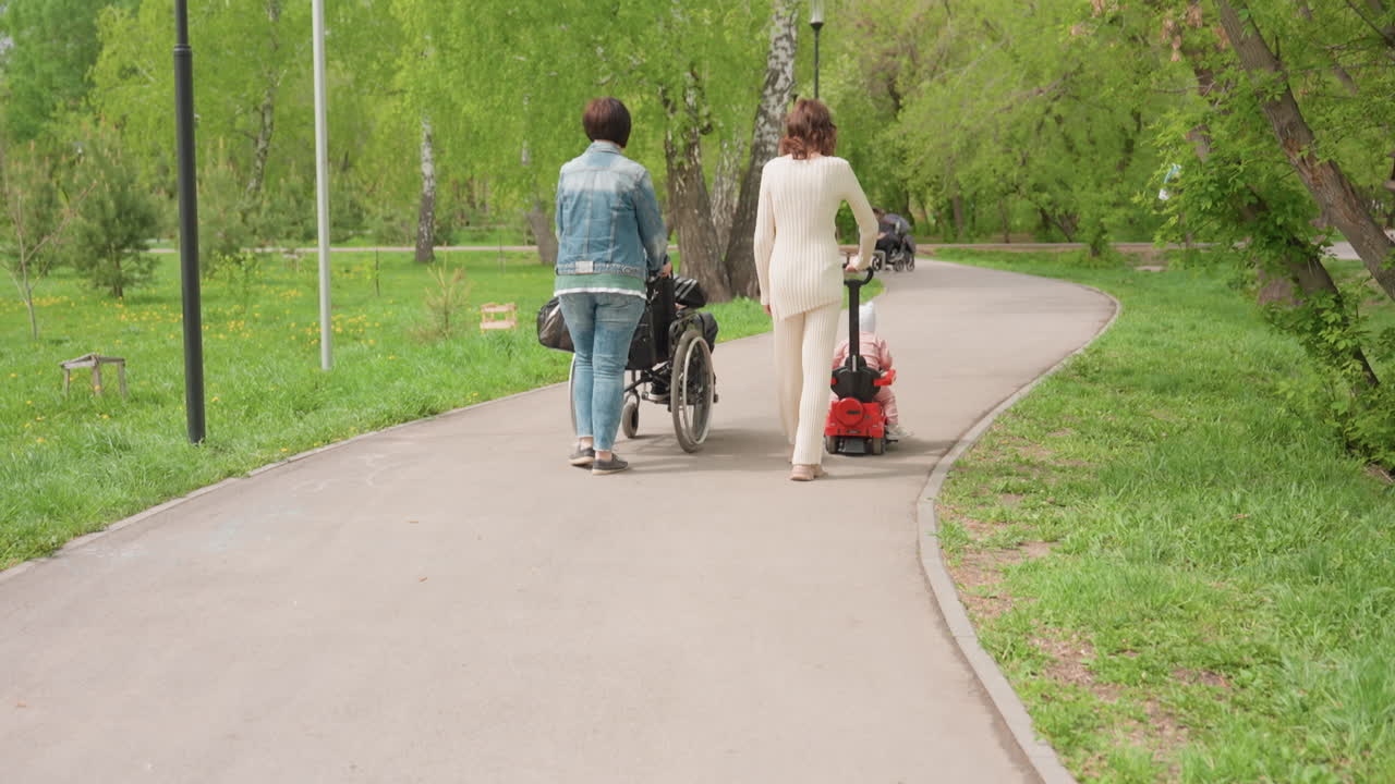 Curved Park Path Caregiver Guiding Wheelchair And Toddler Scooter, Leafy Trees, Smooth Pavement, Inclusive Companionship, Gentle Pace, Urban Green Space, Supportive Gesture, Daytime Stroll