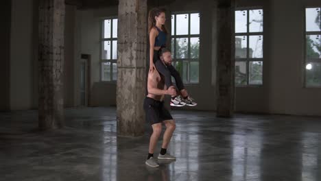 Athletic-shirtless-man-crouches-with-a-girl-on-his-shoulders-in-empty-loft-studio.-Does-squats-with-his-partner-on-shoulders