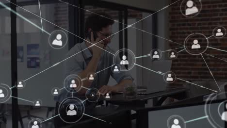 Animation-of-network-of-people-icons-over-man-on-smartphone-working-with-laptop-in-office