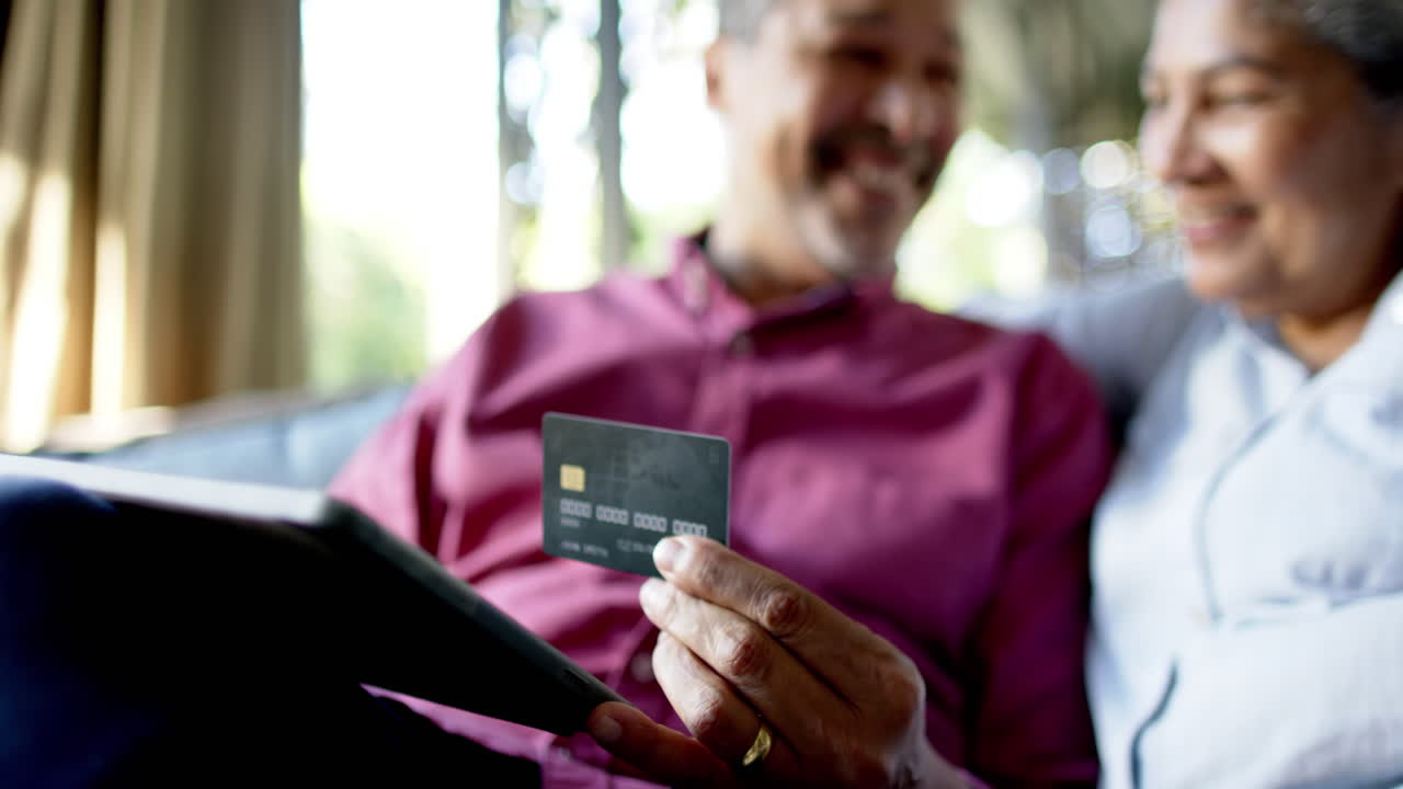 una pareja de ancianos feliz y diversa sentada en el sofá y usando una tableta para comprar en línea en casa