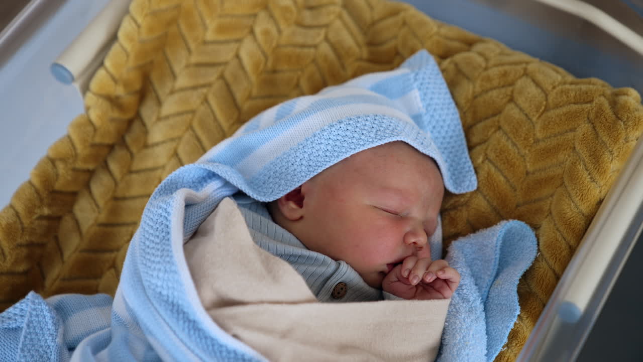 Cute sleeping newborn baby boy lies in a crib. Child in blue blanket holding tiny hand near the mouth. Top view.