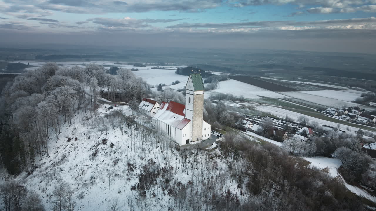 Un drone inizia una seconda orbita attorno alla chiesa bianca innevata con il tetto rosso e agli edifici circostanti sulla cima della collina a Utterweiler, Baden-Württemberg, tra alberi gelidi