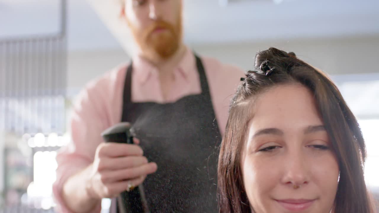 Caucasian male hairdresser spraying happy female client's hair with water at salon, slow motion